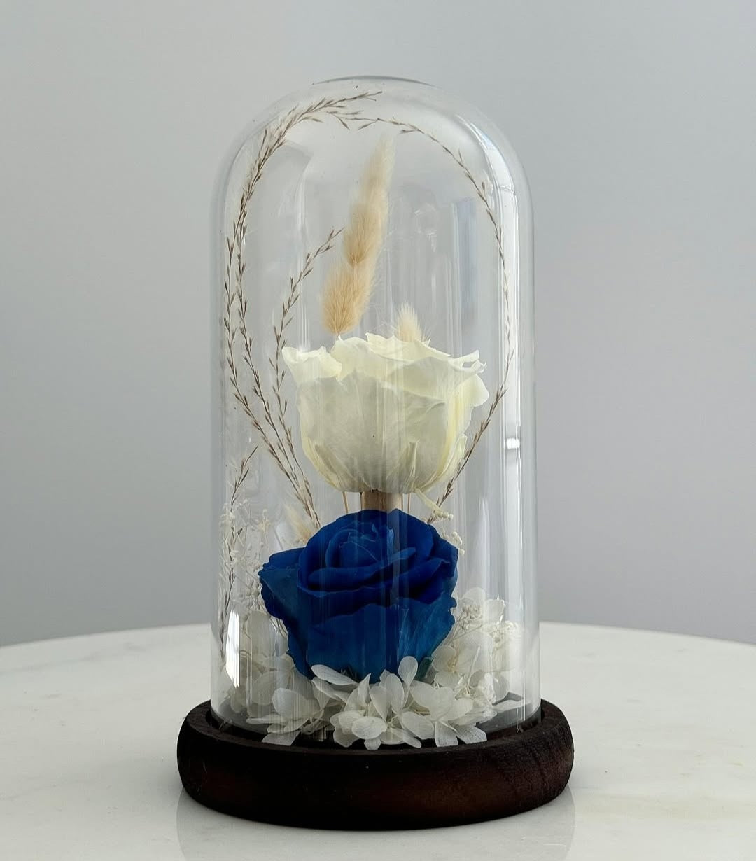 "Elegant preserved flower arrangement in a glass dome with a dark wooden base. Features a striking blue rose beneath a delicate white rose, accented with dried floral elements for a timeless and sophisticated display. Perfect for home décor, gifts, or special occasions."